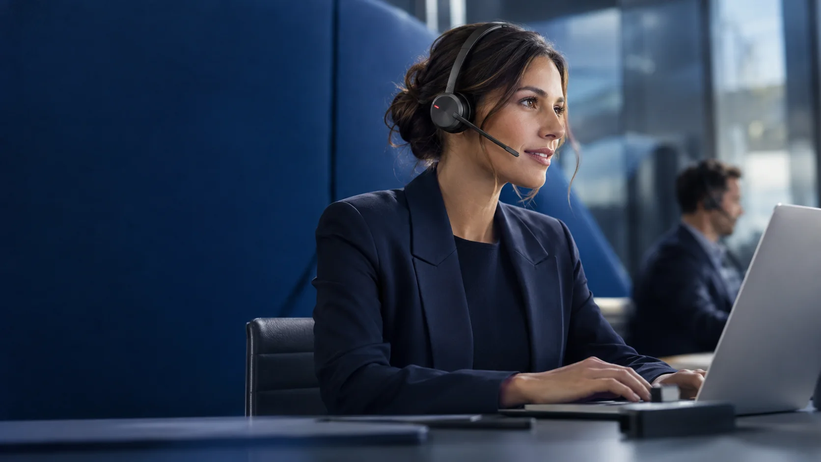 A woman wearing a headset works at a laptop in a bright office with navy panels, while a colleague on a headset works in the background.